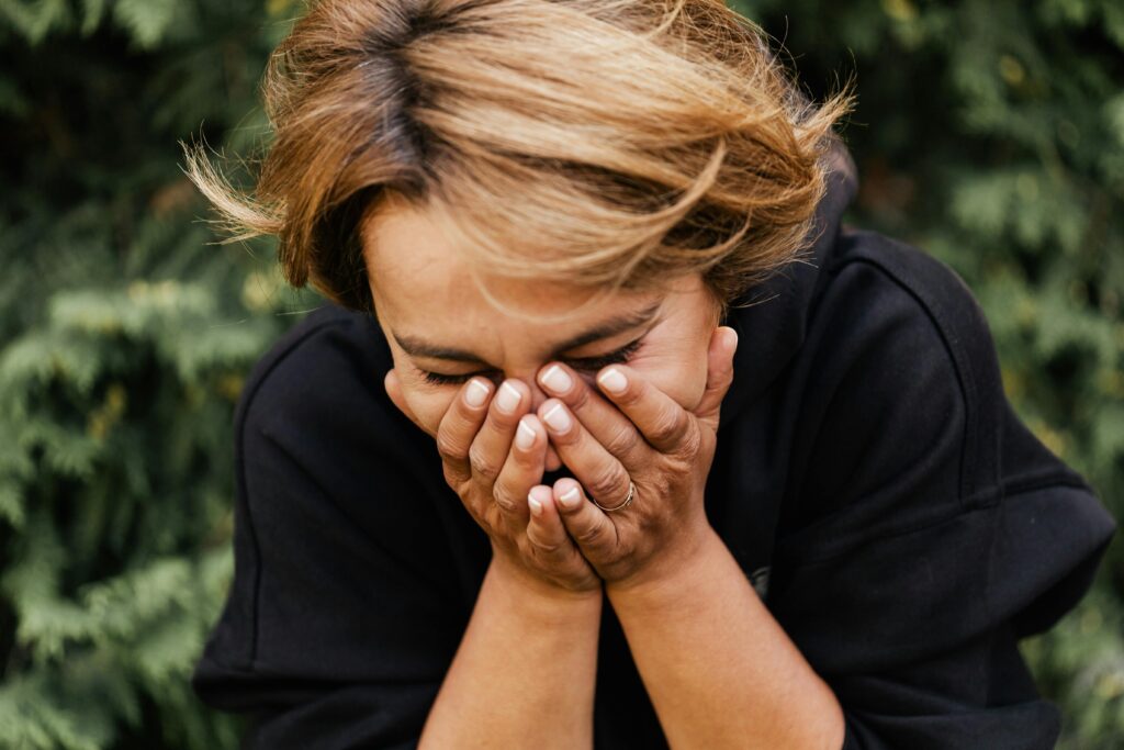 A woman outdoors with hands covering her face, expressing emotion and introspection.