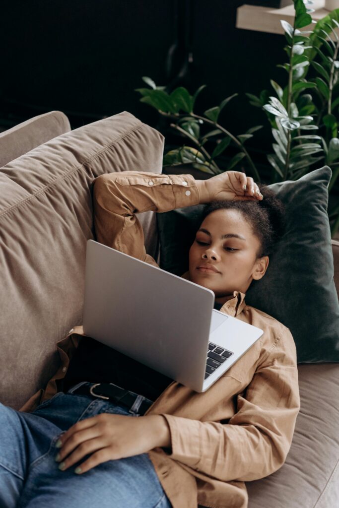 African American woman lying on a couch using a laptop, surrounded by lush indoor plants.