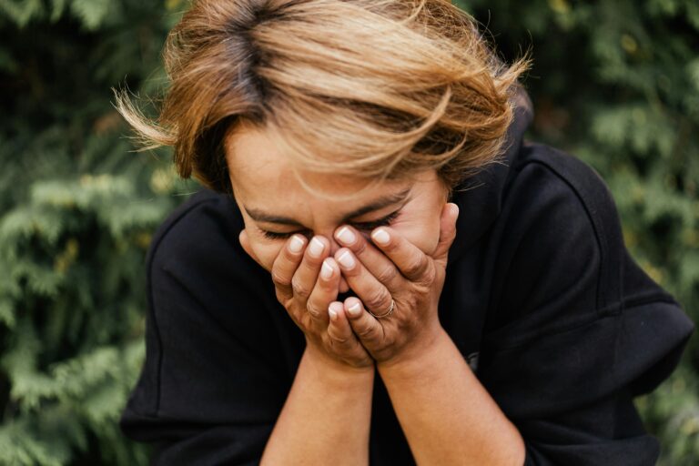 A woman outdoors with hands covering her face, expressing emotion and introspection.