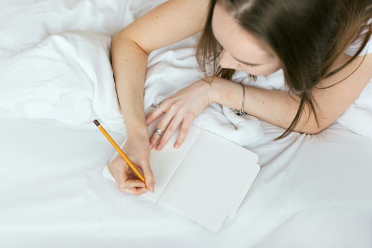 Woman relaxing on a bed while writing in a journal with a pencil.