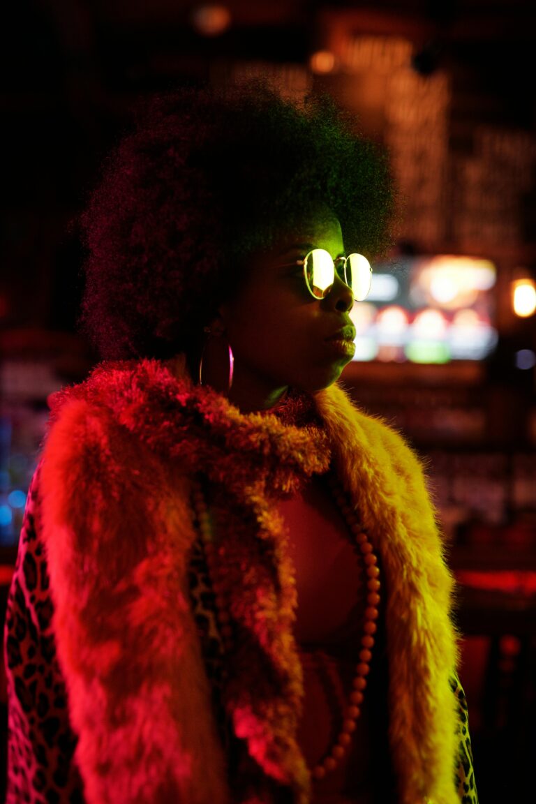 Portrait of a stylish woman in sunglasses and fur coat in a dimly lit nightclub with colorful lights.